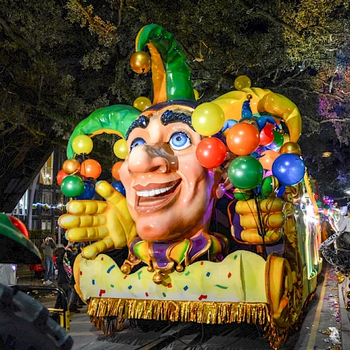 A colorful parade float with a jester face and balloons is being photographed by a crowd under night lights.