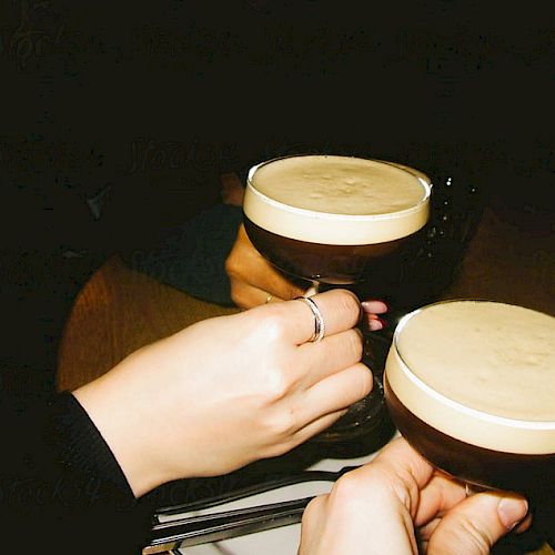 Two people clinking glasses filled with a creamy, frothy drink in a dimly lit setting.