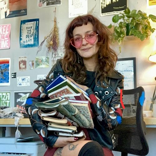 A person with wavy hair and pink-tinted sunglasses sits in an office, holding a stack of colorful notebooks and magazines in front of a wall filled with posters and plants.