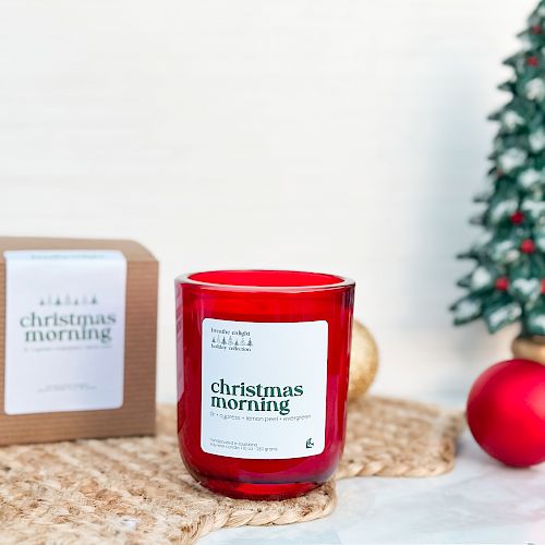 A red scented candle in a glass jar sits on a woven mat with a small “Christmas morning” label, a gift box, and a Christmas tree ornament in the background.