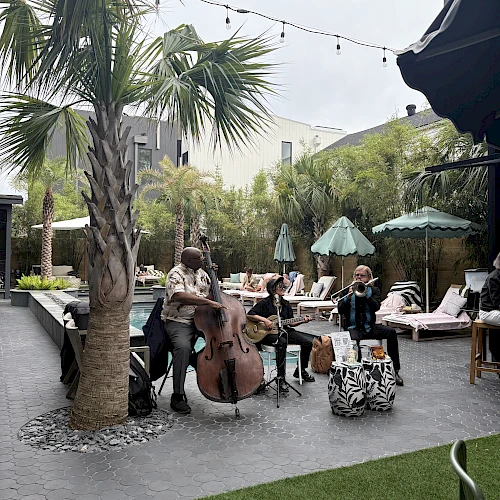 A small live band plays by palm trees at an outdoor patio, with string lights, umbrellas, and lounge seating in the background.