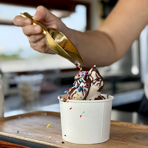 A hand drizzles melted chocolate and colorful sprinkles onto a soft-serve ice cream in a cup, using a gold-tinted spoon.