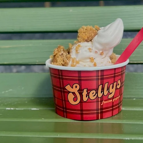 A red plaid ice cream cup labeled “Stella” sits on a green bench, topped with soft-serve and crunchy toppings, with a red spoon.