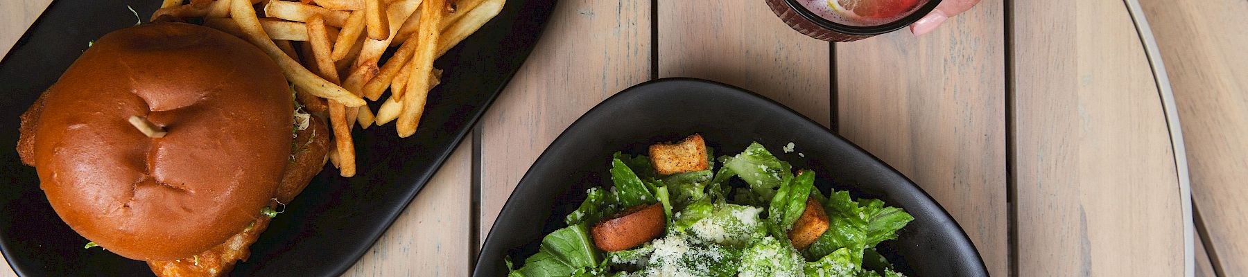 Burger and fries, Caesar salad, berry soda in a glass, and a bowl of roasted vegetables on the table.