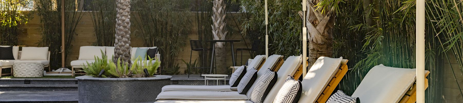 Poolside patio with white lounge beds under green striped umbrellas, pillows, and a small pool fountain area nearby.