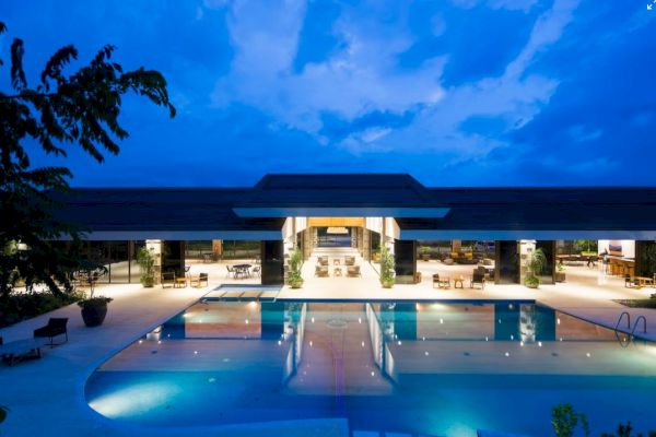 A luxurious modern building with an illuminated pool in the foreground, surrounded by outdoor seating, under a twilight sky.