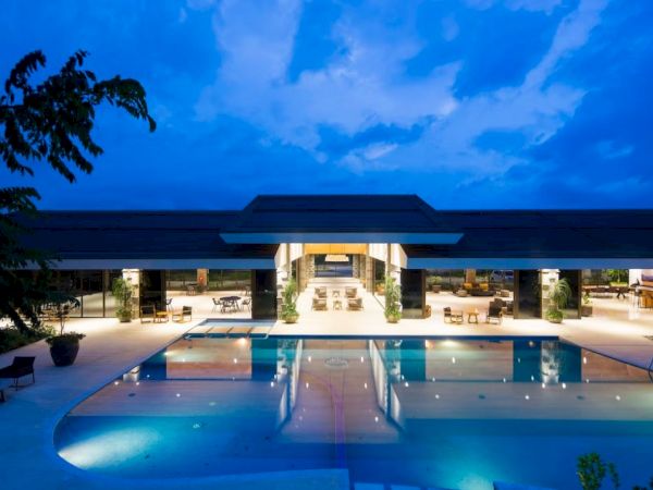 A luxurious modern building with an illuminated pool in the foreground, surrounded by outdoor seating, under a twilight sky.