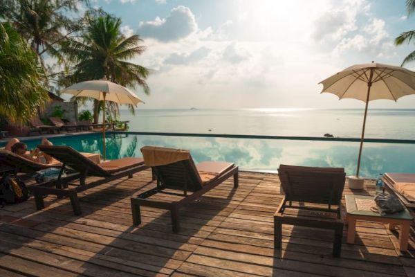 A serene poolside scene with sun loungers, umbrellas, palm trees, and a calm ocean view under a partly cloudy sky.