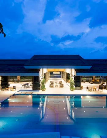 A luxurious building with a large, illuminated swimming pool and lounge area, set against a vibrant evening sky.