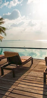 A serene poolside deck with sun loungers and umbrellas facing an infinity pool under a bright sky, surrounded by palm trees.