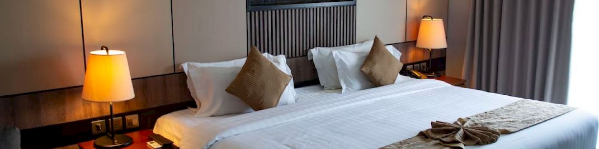 A hotel room with a large bed, two bedside lamps, pillows, a folded towel on the bed, and a window with closed curtains.