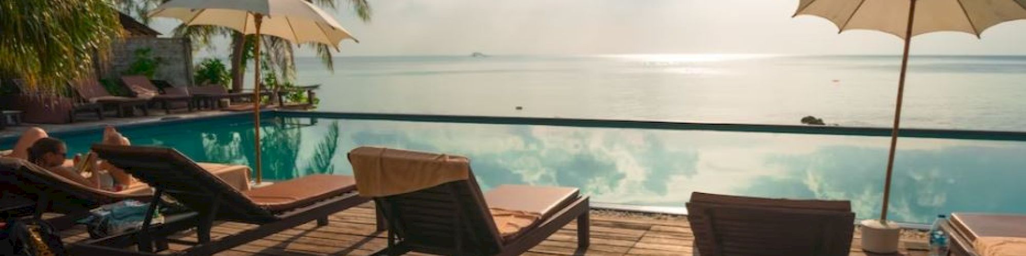 A serene poolside scene with sun loungers, umbrellas, palm trees, and an ocean view under a bright sky.