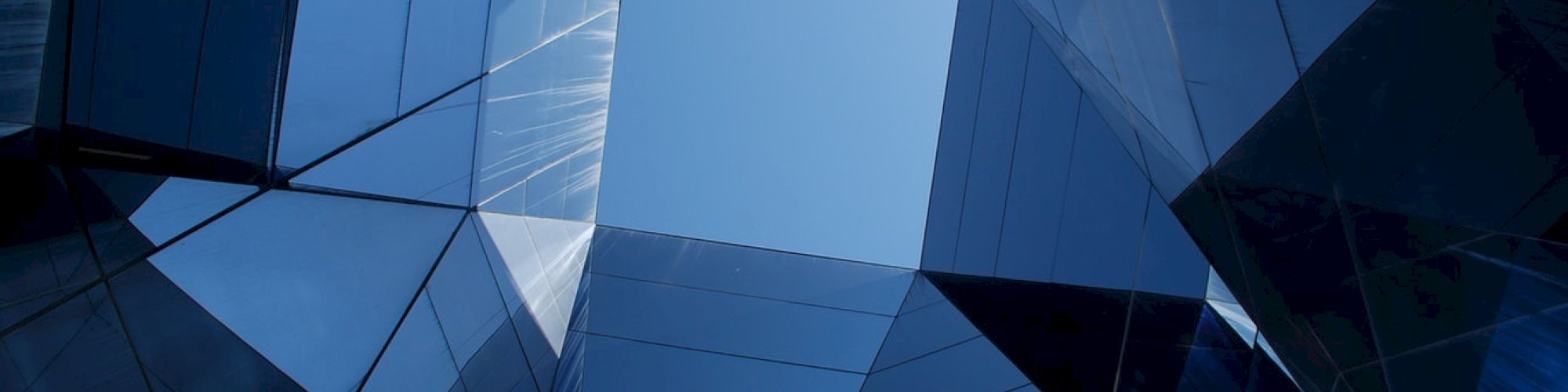 The image shows a geometric view looking up between modern glass buildings, revealing a clear blue sky in the center.