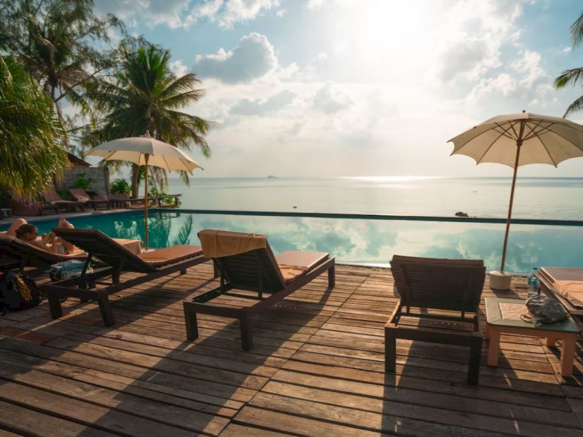 A serene poolside scene with lounge chairs, umbrellas, and a view of the ocean, under a partly cloudy sky with sunlight reflecting on water.
