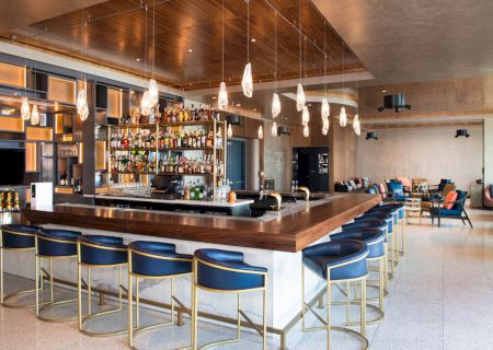 The image shows a modern bar with hanging lights, a stocked drink shelf, and blue cushioned bar stools, set in a sophisticated lounge area.