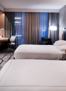 A modern hotel room with two beds, a wall-mounted TV, desk, chair, and patterned carpet, featuring ambient lighting and large curtains.