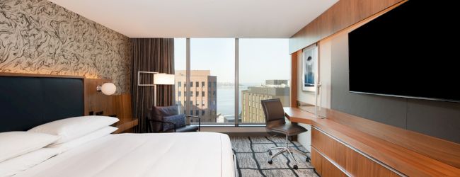 A modern hotel room with a bed, TV, desk, and view of city buildings through large windows, featuring stylish wood and patterned decor.