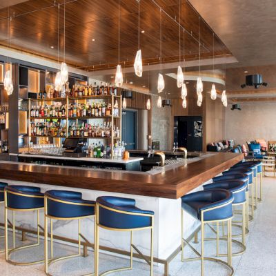 The image shows a modern bar area with a wooden counter, blue stools, hanging lights, and a fully stocked bar shelf in a spacious lounge.