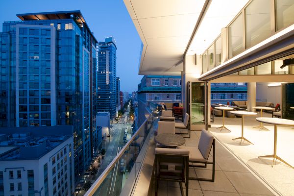 A modern cityscape at dusk with high-rise buildings and a stylish outdoor balcony lounge featuring tables and seating.