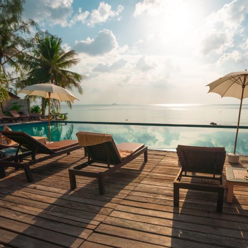 Sun loungers by an infinity pool with umbrellas, overlooking the ocean, surrounded by palm trees under a bright, partly cloudy sky.