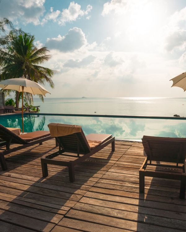 Sun loungers by an infinity pool with umbrellas, overlooking the ocean, surrounded by palm trees under a bright, partly cloudy sky.