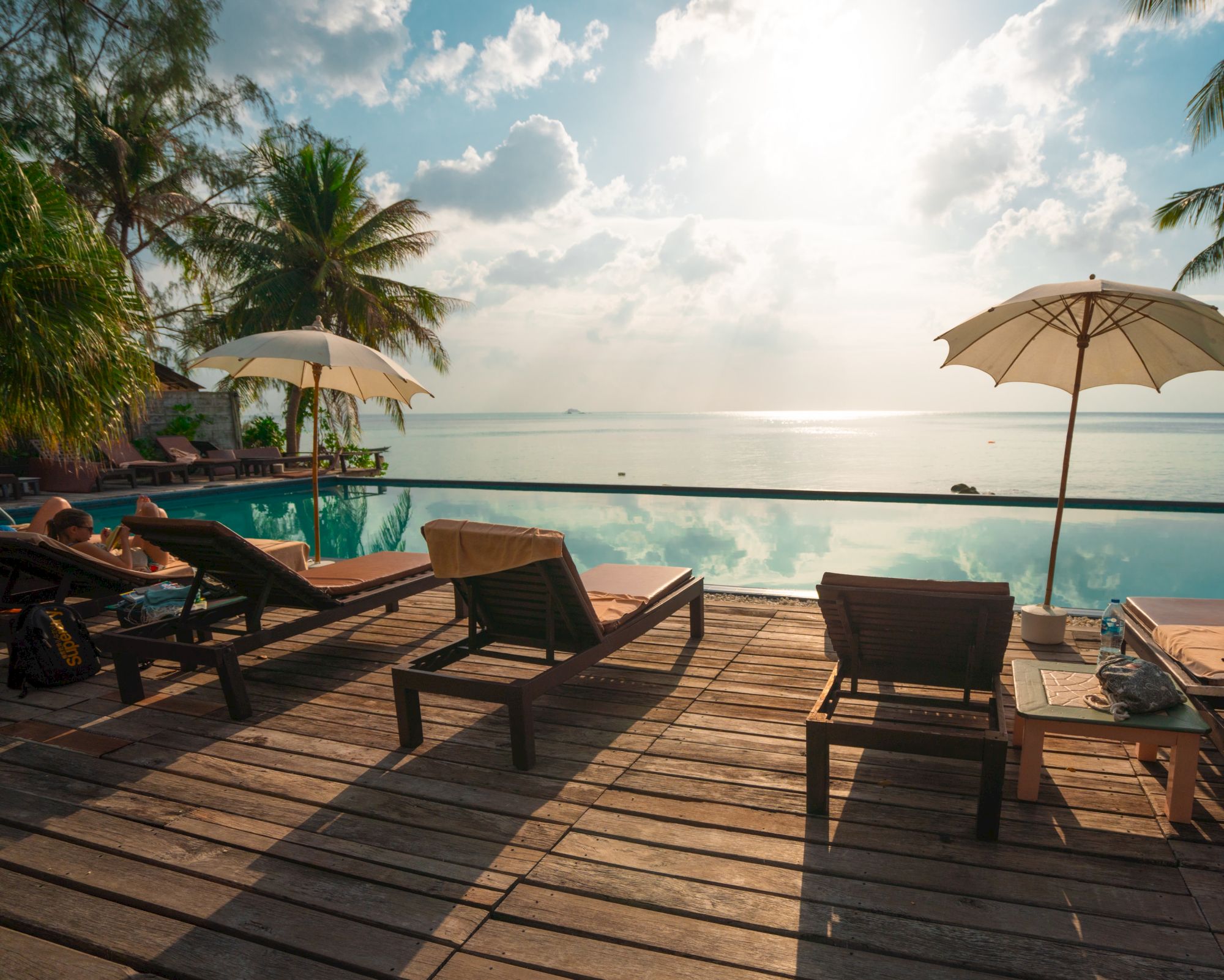 A tropical setting with lounge chairs, umbrellas, and a pool overlooking a calm sea, framed by palm trees under a cloudy blue sky.