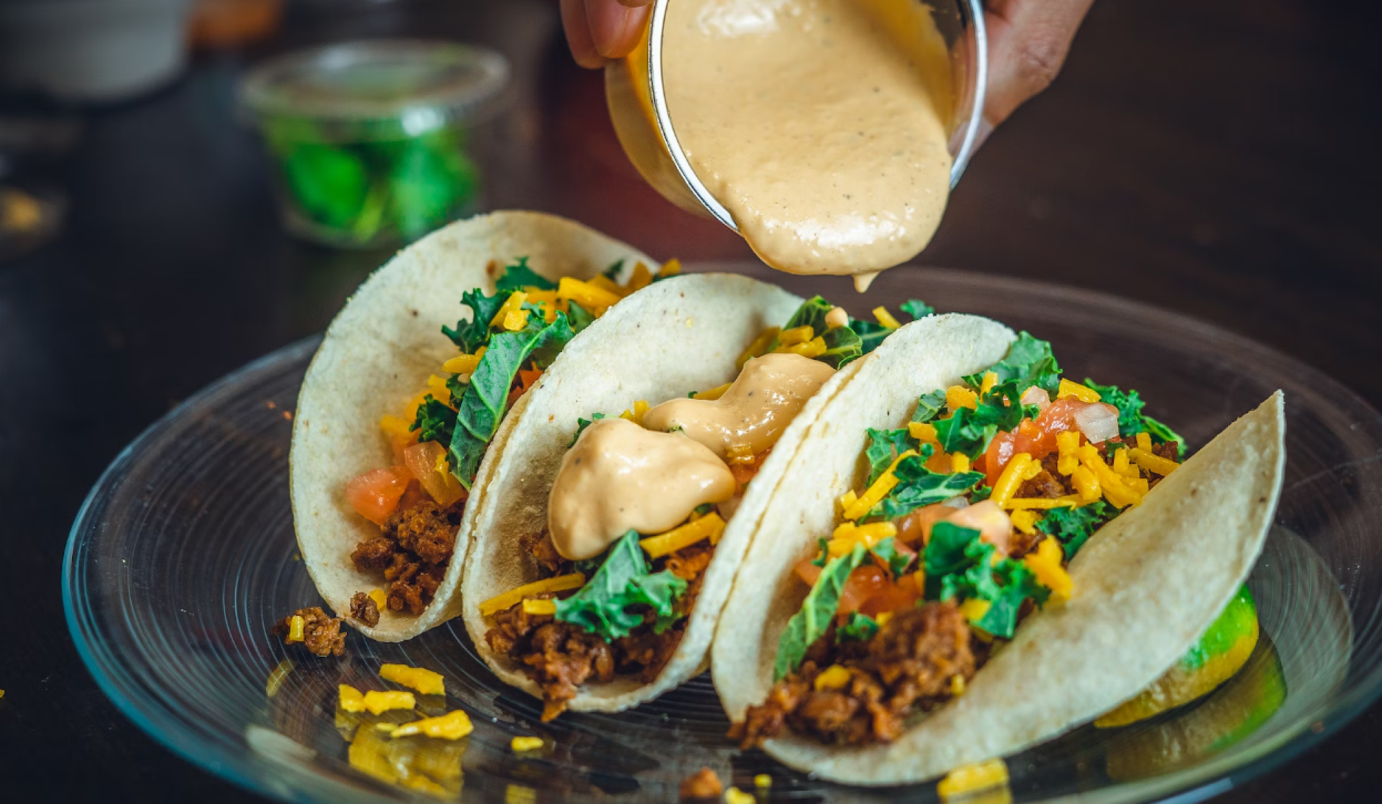 Three tacos filled with meat, cheese, lettuce, and tomatoes on a plate, with sauce being poured over them.