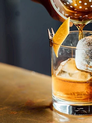 A drink is being poured into a glass with ice and a lemon twist garnish on a wooden surface.
