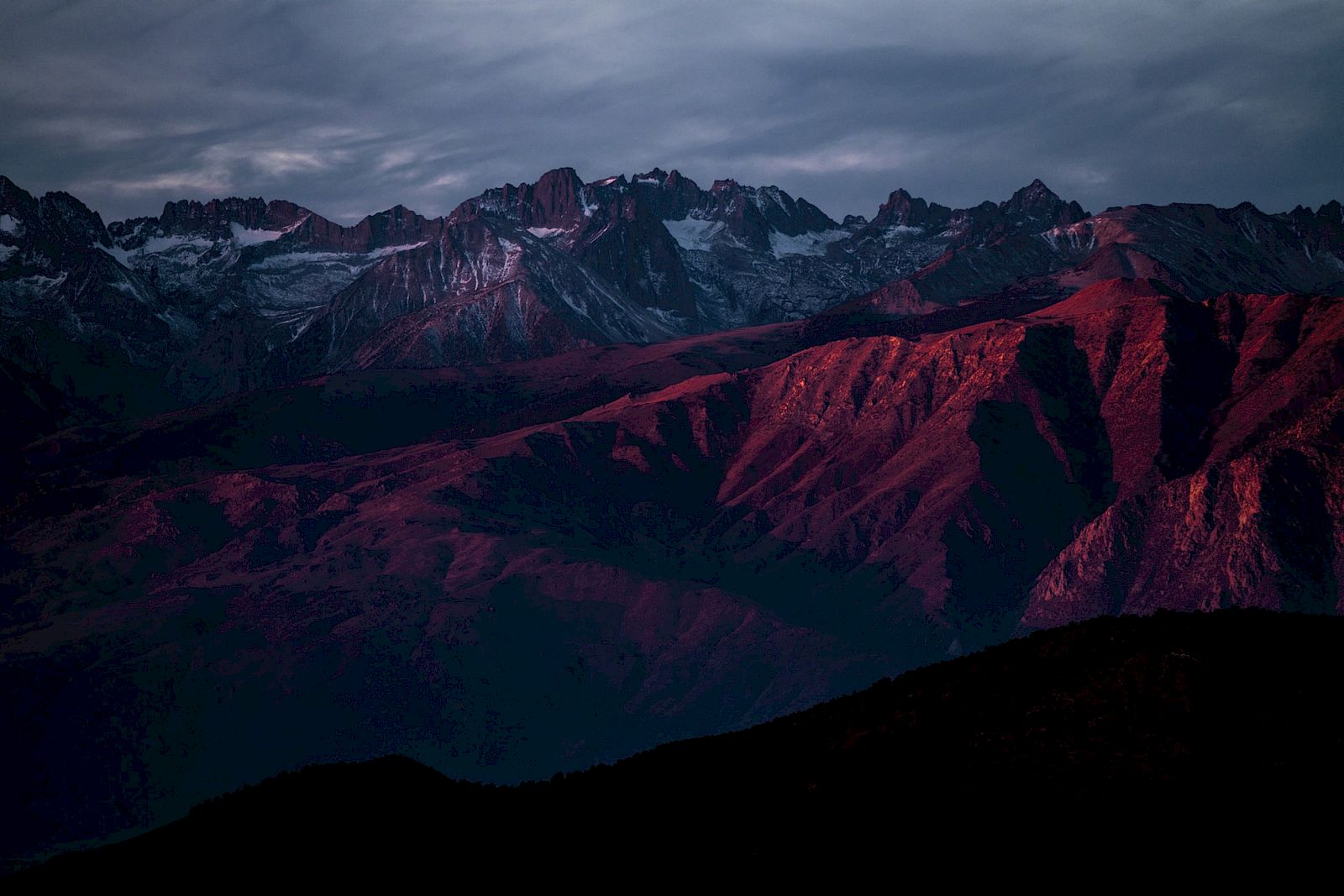 A mountain range with snow-capped peaks bathed in a red sunset glow under a cloudy sky.
