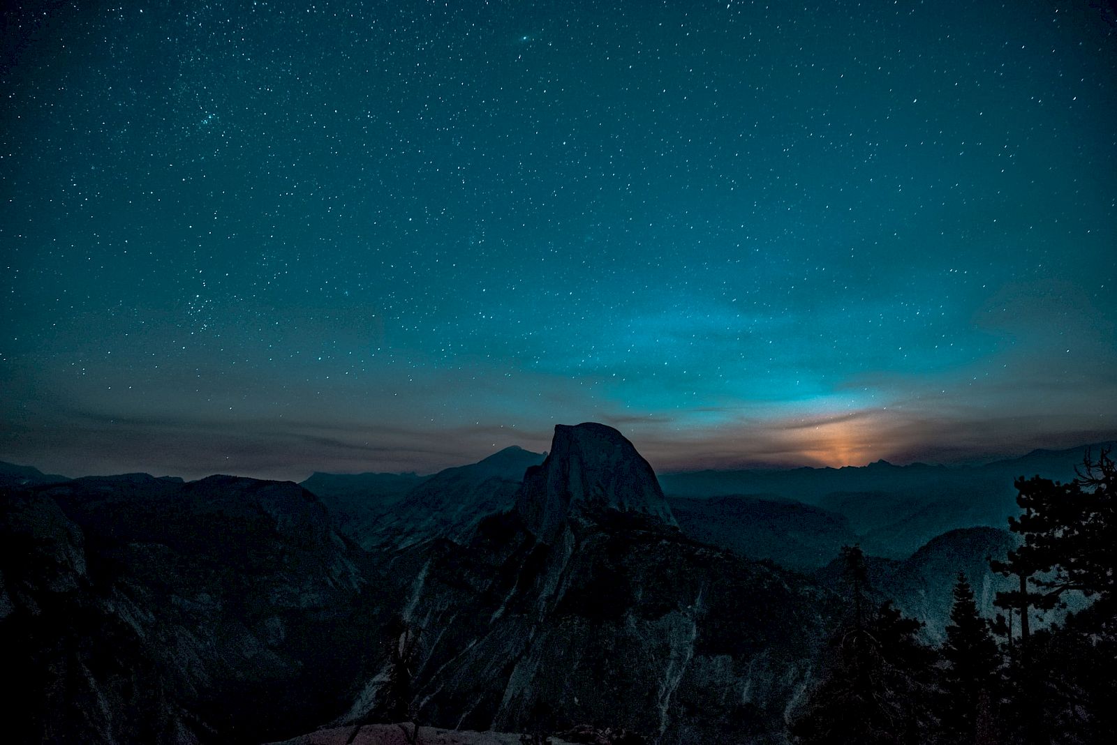 A night sky filled with stars is above a mountain silhouette, with a subtle glow on the horizon, creating a serene landscape scene.