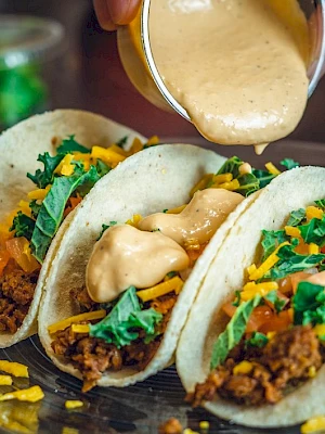 Three tacos with ground meat, cheese, lettuce, and tomatoes are on a plate, being topped with a creamy sauce.