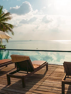 Poolside scene with lounge chairs, umbrellas, and palm trees overlooking a calm ocean and sky with clouds, bathed in sunlight.