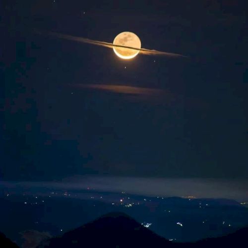 A full moon appears with two thin clouds encircling it, resembling Saturn's rings, above a landscape with scattered lights below.