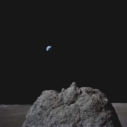 A lunar surface landscape with a large rock in the foreground and the Earth visible in the dark sky above.
