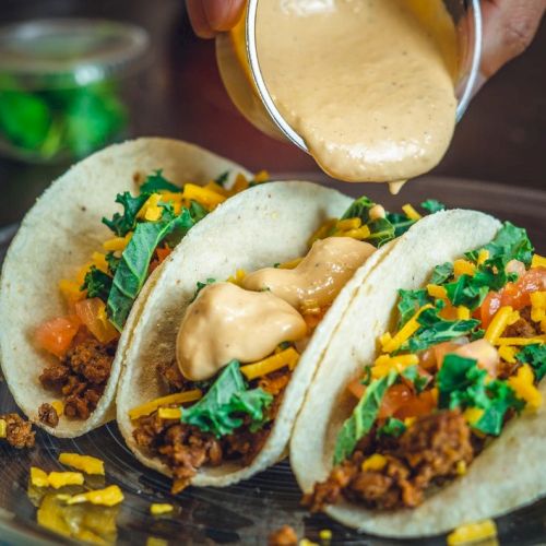 Three tacos on a plate, filled with meat, cheese, lettuce, and diced tomatoes, with sauce being poured over them.