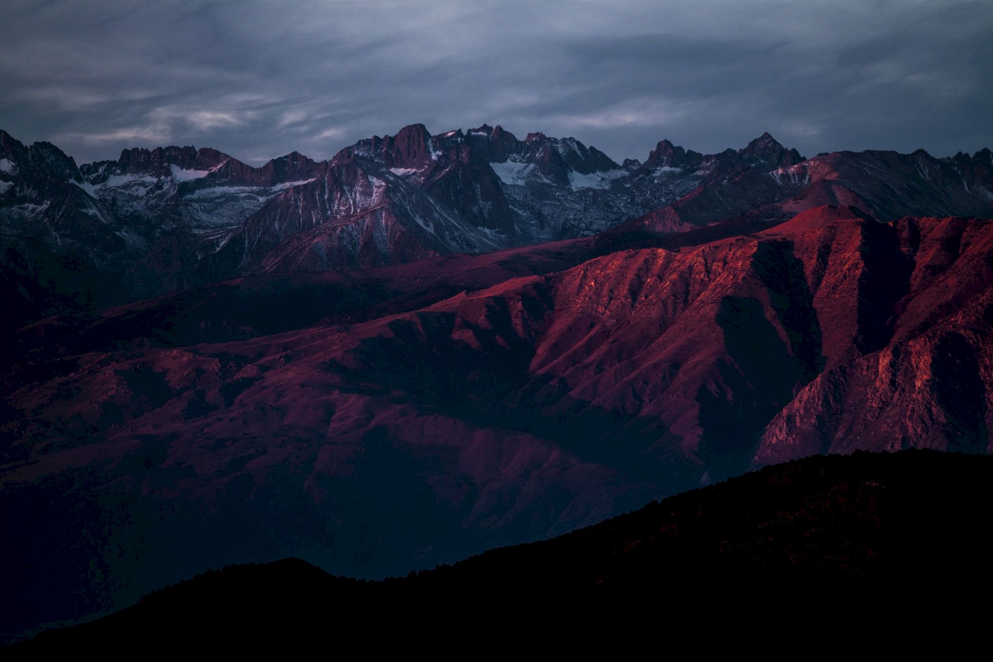 A rugged mountain range with peaks partly covered in snow, bathed in red and purple hues under a dramatic, cloudy sky.