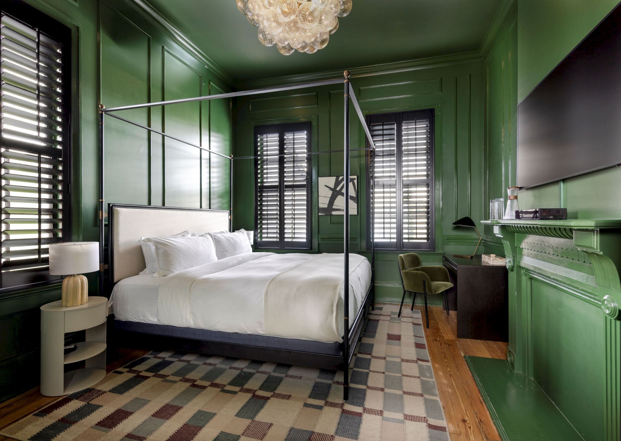 A bedroom with green walls features a four-poster bed, lamp, and TV, enhancing its classic look with modern and cozy décor elements.