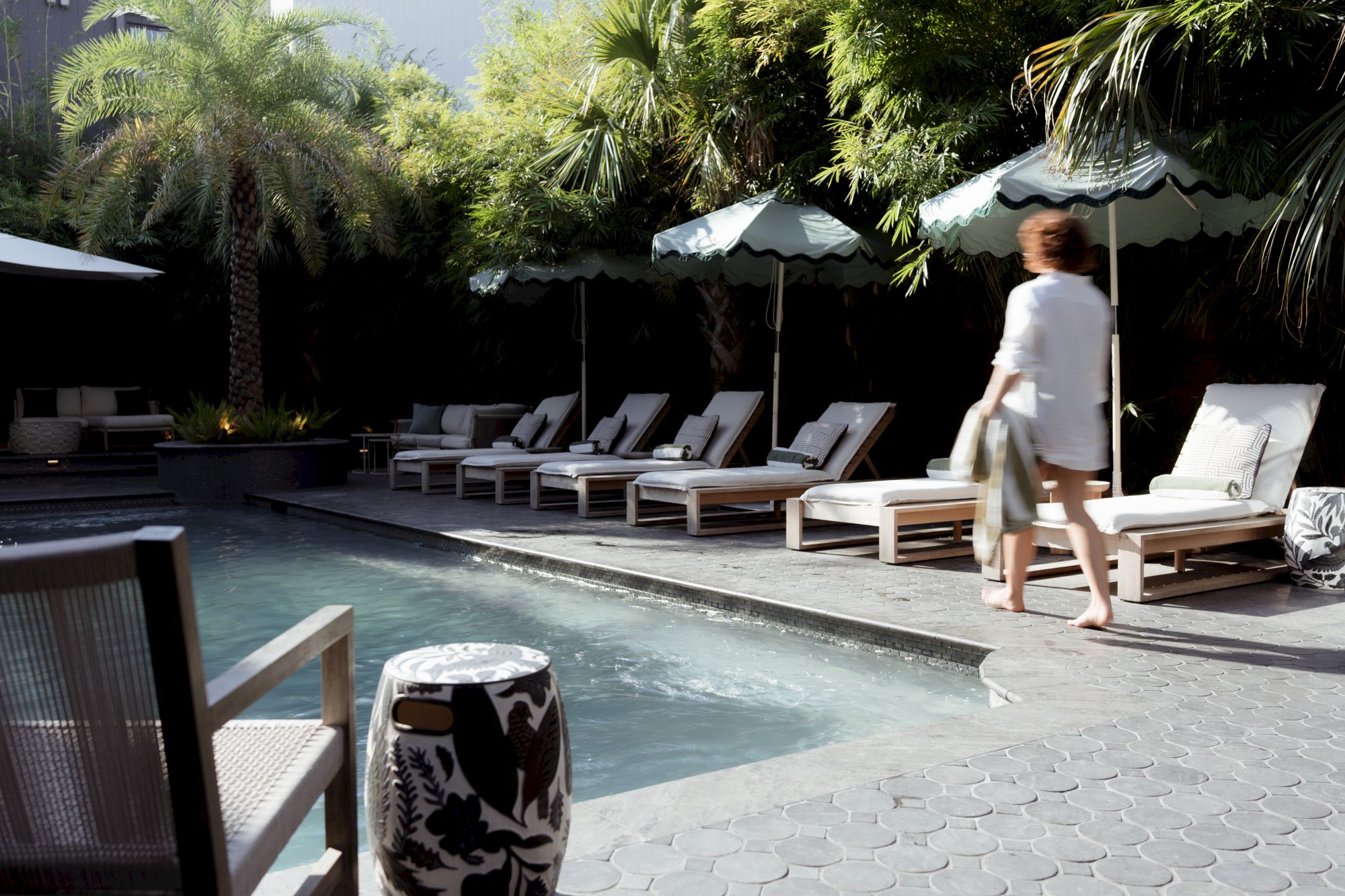 A person walks by a sunny pool area with lounge chairs, umbrellas, and palm trees in the background.