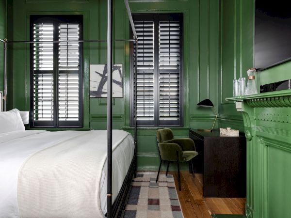 A green-paneled room with a bed, desk, chair, and shutters. Modern decor with a rug, lamp, and wall art.