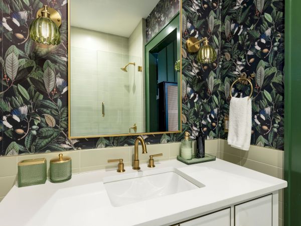 The image shows a bathroom with floral wallpaper, a white sink, brass fixtures, and green accents, including a towel and candles.