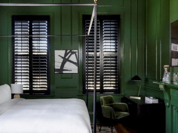 A stylish bedroom with green walls, a canopy bed, patterned rug, modern chandelier, and shuttered windows, creating a cozy ambiance.