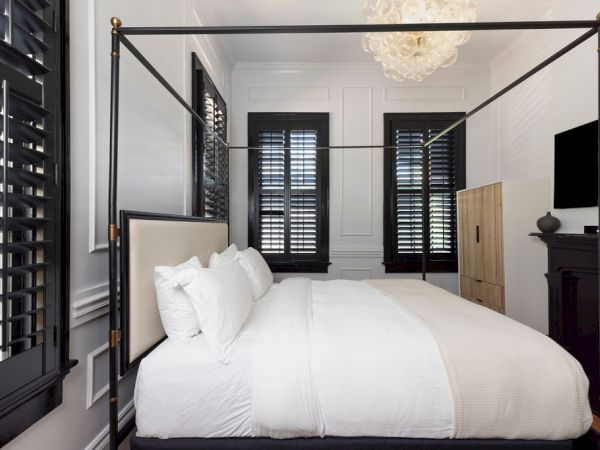 The image shows a bedroom with a four-poster bed, white bedding, black window shutters, a chandelier, and a wall-mounted TV.
