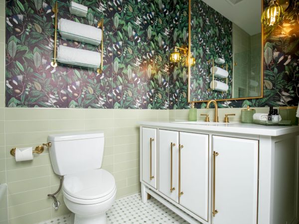 A bathroom with floral wallpaper, white cabinets, double sinks, a toilet, and gold fixtures. Towel rack and wall lights enhance decor.