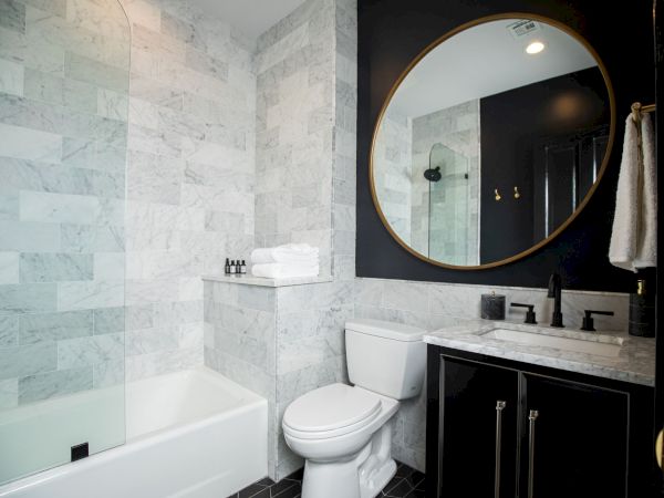 A modern bathroom features a bathtub, toilet, and vanity with a large round mirror, set against marble and black accents.