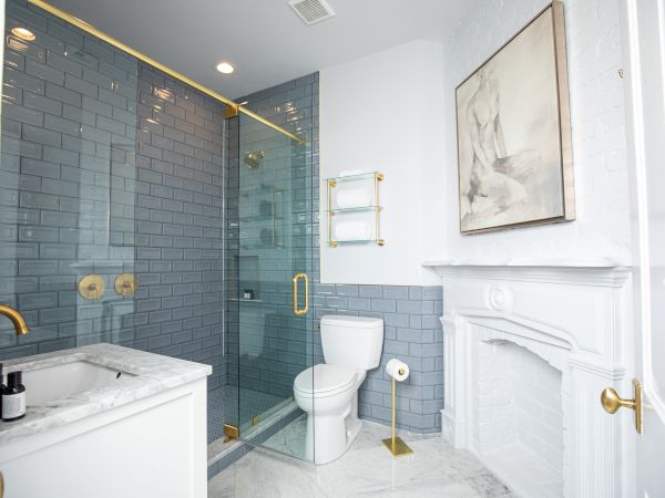 The image shows a modern bathroom with a glass shower, marble sink, toilet, and wall art, featuring gold fixtures and gray tiles.