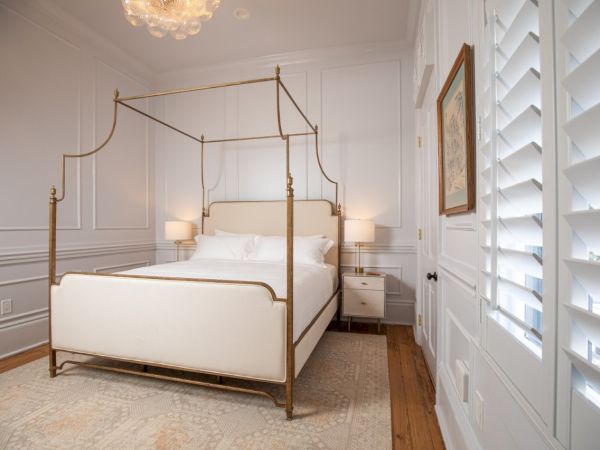 A bedroom with a canopy bed, white bedding, two nightstands with lamps, a rug, a framed picture, and shutters on the windows.