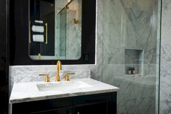 A luxurious bathroom with marble surfaces, gold fixtures, a mirror, glass shower, and neatly displayed toiletries on a shelf.