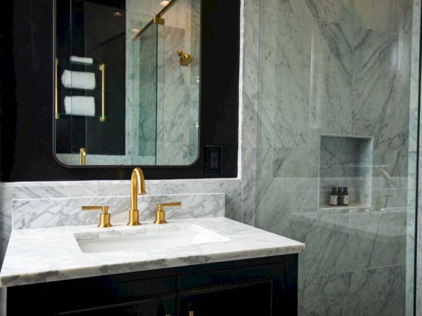 A luxurious bathroom with marble surfaces, gold fixtures, a mirror, glass shower, and neatly displayed toiletries on a shelf.