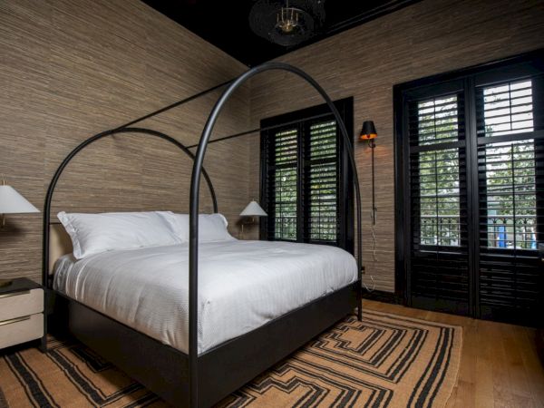 A bedroom with a canopy bed, white bedding, neutral walls, wooden shutters, lamps, and a striped rug on a wooden floor.