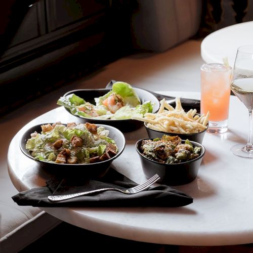 A table set with salad, fries, and drinks, including a glass of wine and a pink cocktail, inviting a cozy dining experience.